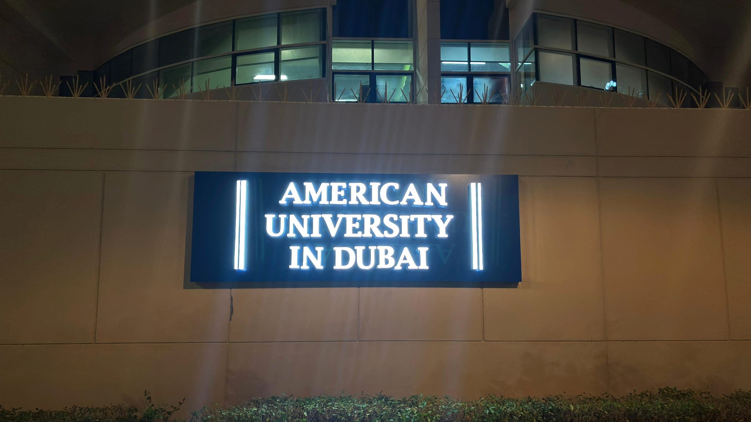 Illuminated American University in Dubai sign glowing on a boundary wall at night