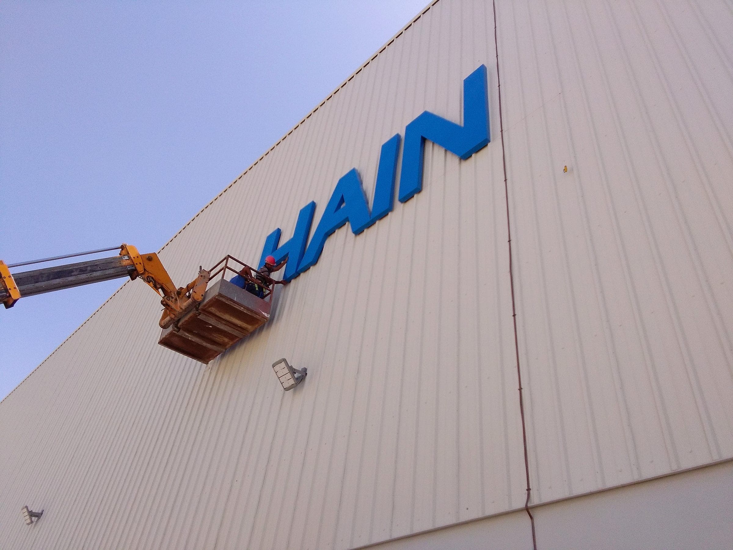 Installers on a boom lift fixing blue HAIN channel letters to a factory façade